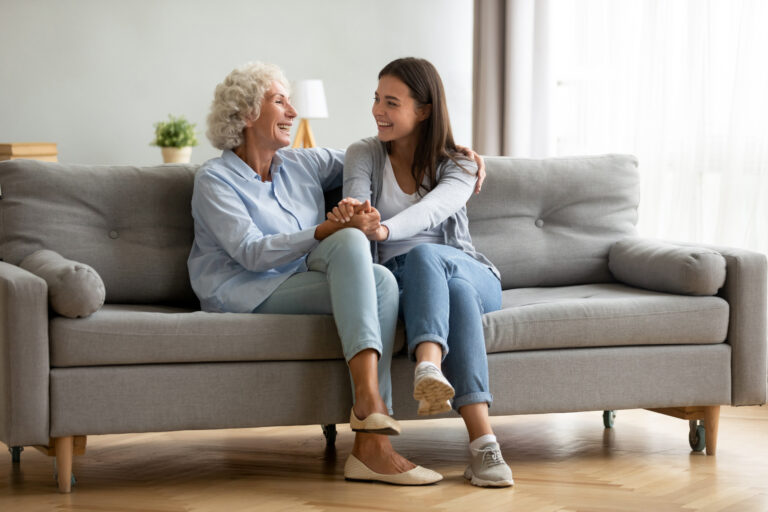 Granddaughter and teenage granddaughter sitting on couch together smiling at one another - Long-distance teenage grandchildren - The Grandkid Connection