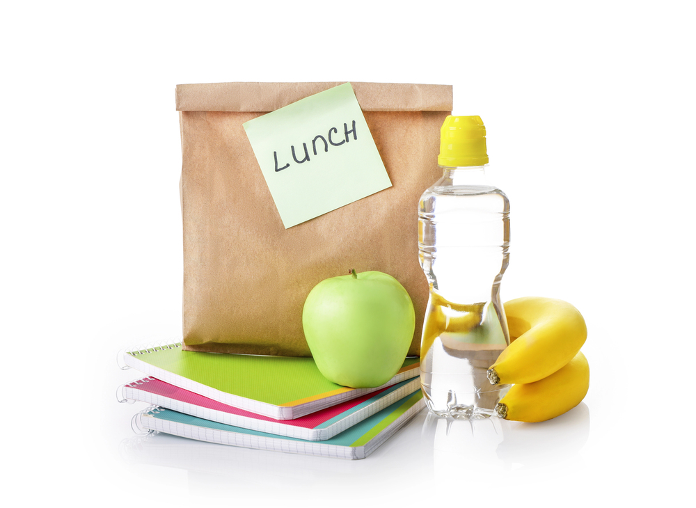 A brown lunch bag sitting on top of colorful notebooks along with an apple, a bottle of water, and two bananas - The Grandkid Connection